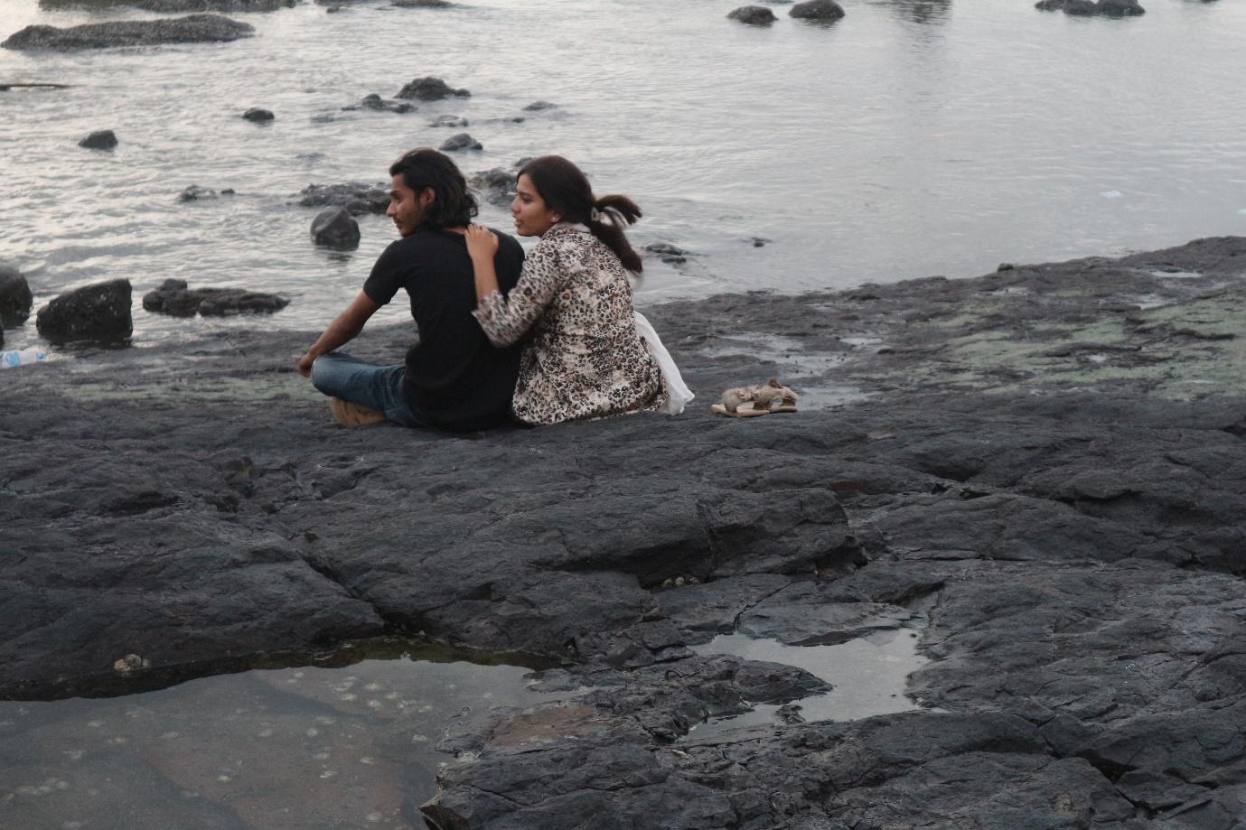 A man and woman are seated close to each other; both their heads are turned towards the left; the woman’s arm rests on the man’s back, her fingers slipping into the neck of his t-shirt; her slippers are placed neatly next to her and she seems to be mid-sentence.